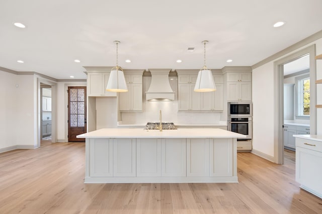 kitchen featuring premium range hood, an island with sink, decorative light fixtures, and light wood-type flooring