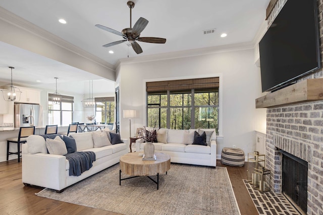 living room featuring crown molding, dark wood-type flooring, ceiling fan with notable chandelier, and a brick fireplace