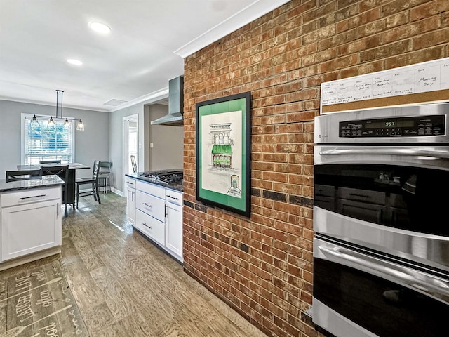 kitchen featuring wall chimney exhaust hood, hanging light fixtures, dark stone counters, light hardwood / wood-style floors, and appliances with stainless steel finishes