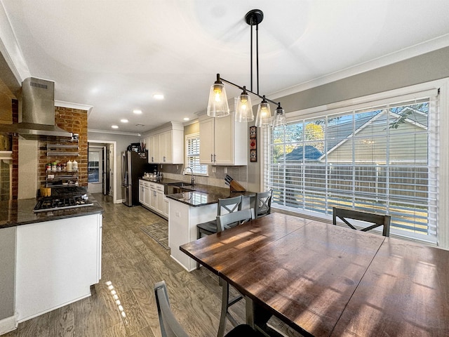 kitchen featuring tasteful backsplash, ornamental molding, wall chimney range hood, white cabinets, and hanging light fixtures