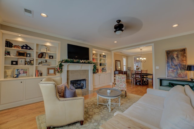 living room featuring a tile fireplace, built in shelves, light hardwood / wood-style floors, ceiling fan with notable chandelier, and ornamental molding