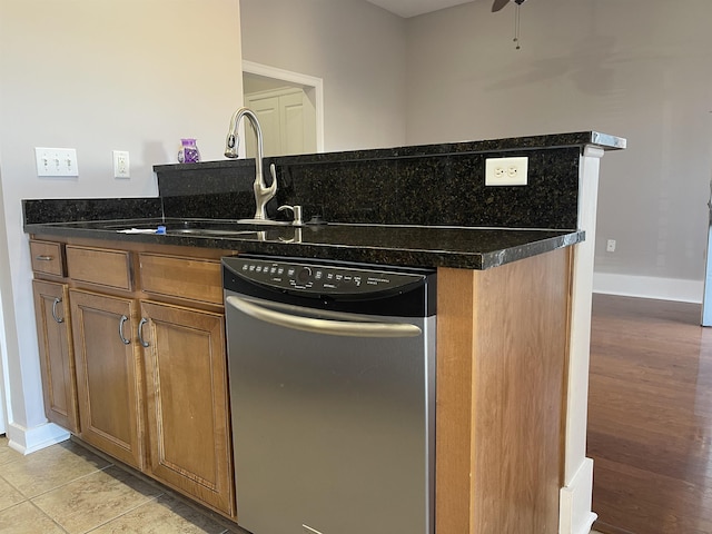 kitchen featuring stainless steel dishwasher, brown cabinetry, light tile patterned flooring, dark stone countertops, and baseboards