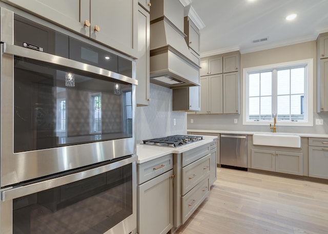 kitchen featuring premium range hood, sink, light wood-type flooring, ornamental molding, and appliances with stainless steel finishes