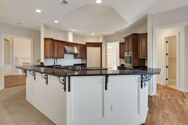 kitchen with kitchen peninsula, a breakfast bar, wall chimney range hood, dark stone countertops, and light hardwood / wood-style floors