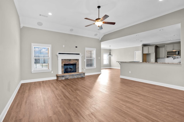 unfurnished living room with wood-type flooring, ceiling fan with notable chandelier, a stone fireplace, and ornamental molding