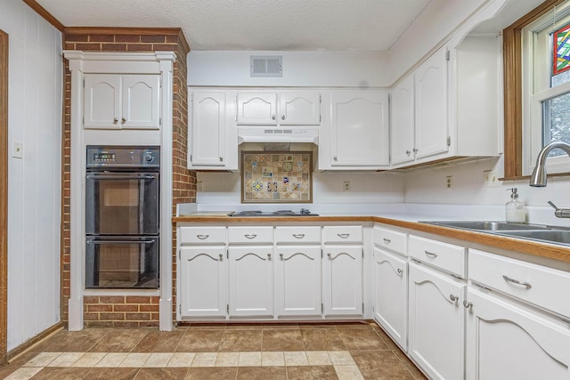 kitchen with stainless steel gas stovetop, sink, black double oven, white cabinetry, and brick wall