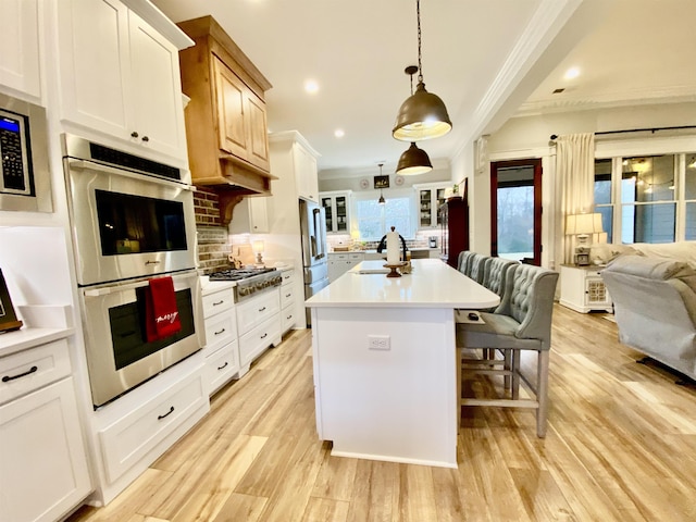 kitchen featuring stainless steel appliances, crown molding, decorative light fixtures, white cabinetry, and an island with sink