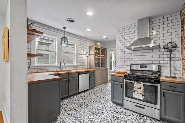 kitchen with butcher block counters, sink, hanging light fixtures, wall chimney range hood, and appliances with stainless steel finishes