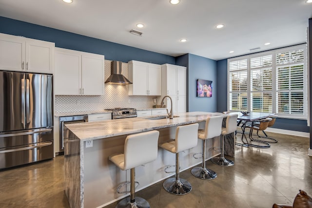 kitchen with wall chimney range hood, sink, appliances with stainless steel finishes, a kitchen island with sink, and white cabinetry