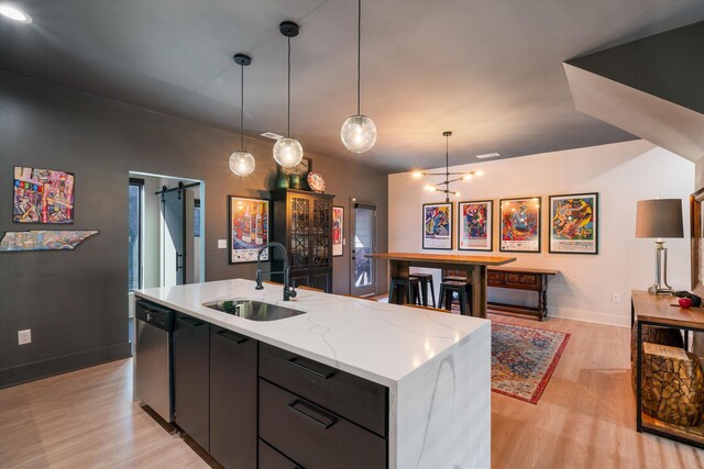 kitchen featuring stainless steel dishwasher, sink, a barn door, hanging light fixtures, and an island with sink