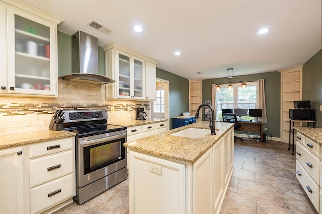 kitchen featuring wall chimney range hood, sink, stainless steel range with electric stovetop, hanging light fixtures, and a center island with sink