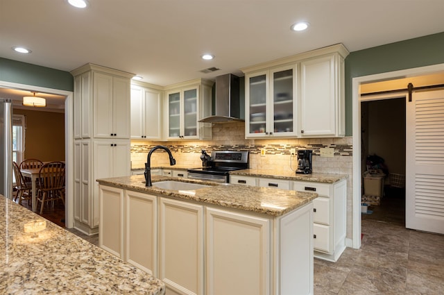 kitchen featuring wall chimney range hood, sink, an island with sink, stainless steel range with electric cooktop, and a barn door