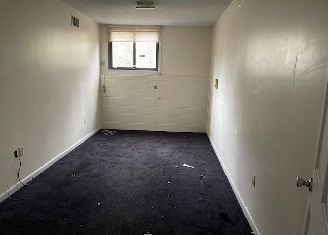 spare room featuring concrete block wall, baseboards, visible vents, a textured ceiling, and dark carpet