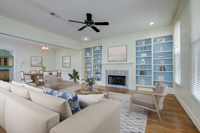 living room featuring built in shelves, dark hardwood / wood-style flooring, crown molding, and ceiling fan with notable chandelier