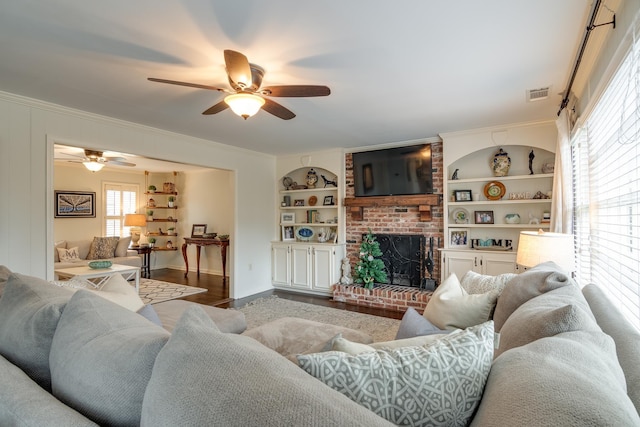 living room with a brick fireplace, built in shelves, ceiling fan, ornamental molding, and wood-type flooring