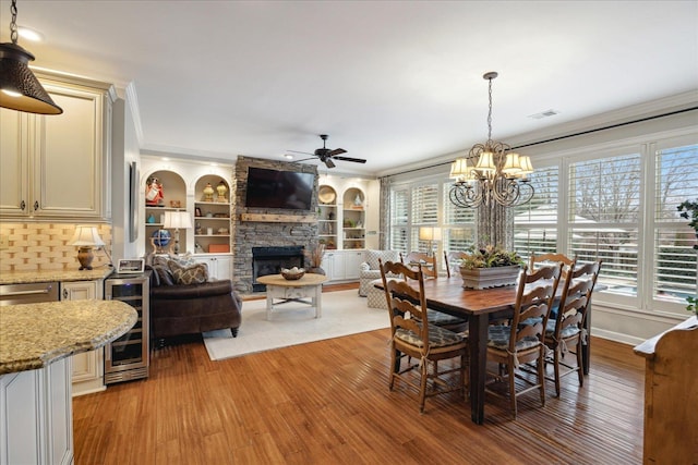 dining space with ceiling fan with notable chandelier, built in features, a fireplace, light hardwood / wood-style floors, and wine cooler