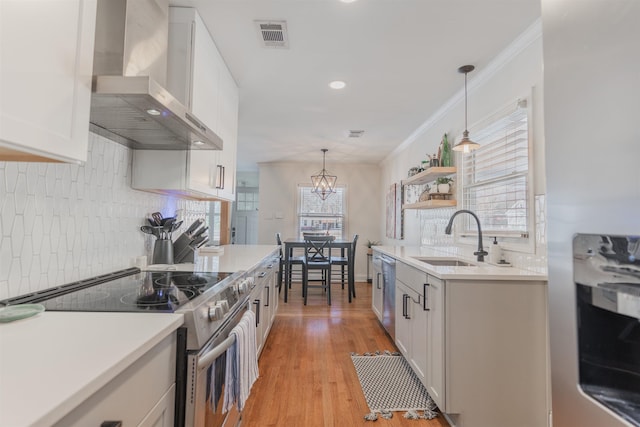 kitchen featuring wall chimney range hood, sink, white cabinetry, hanging light fixtures, and stainless steel appliances