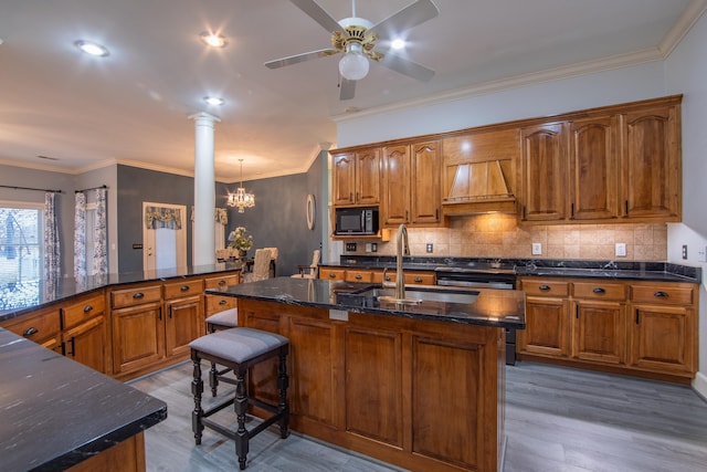 kitchen with black microwave, premium range hood, hardwood / wood-style floors, a kitchen island with sink, and ceiling fan with notable chandelier