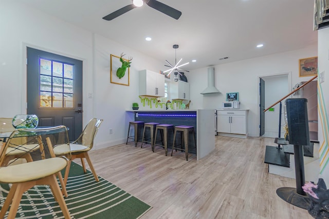 interior space with wall chimney exhaust hood, ceiling fan with notable chandelier, light hardwood / wood-style flooring, white cabinets, and a breakfast bar area