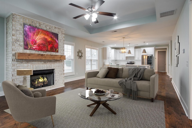 living room featuring a large fireplace, a tray ceiling, ceiling fan, and dark wood-type flooring