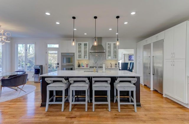 kitchen with appliances with stainless steel finishes, decorative light fixtures, a kitchen island with sink, and wall chimney exhaust hood