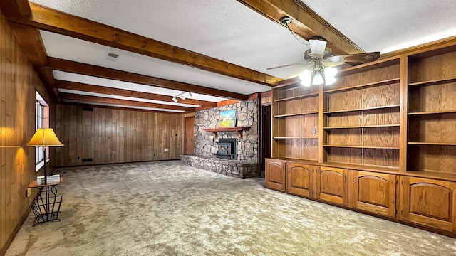 unfurnished living room featuring ceiling fan, wooden walls, beam ceiling, carpet floors, and a wood stove