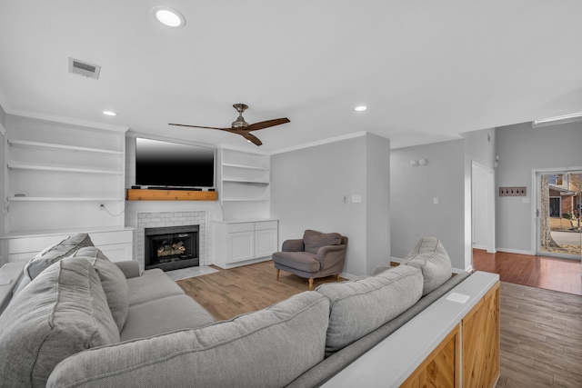 living room with a brick fireplace, built in features, ceiling fan, and light wood-type flooring