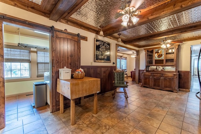 dining area with ceiling fan, a barn door, wooden walls, and beamed ceiling