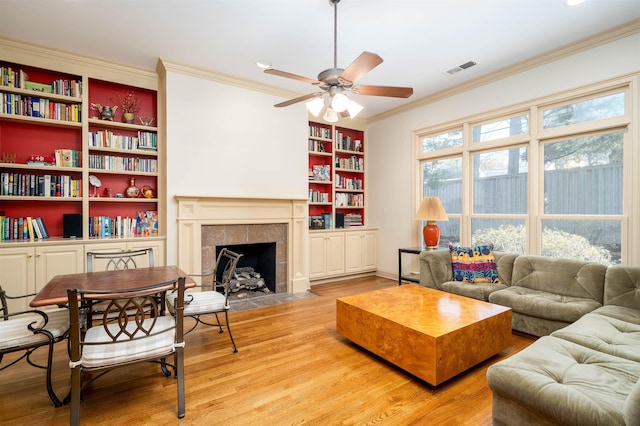 living room with ceiling fan, hardwood / wood-style floors, a tiled fireplace, built in features, and ornamental molding