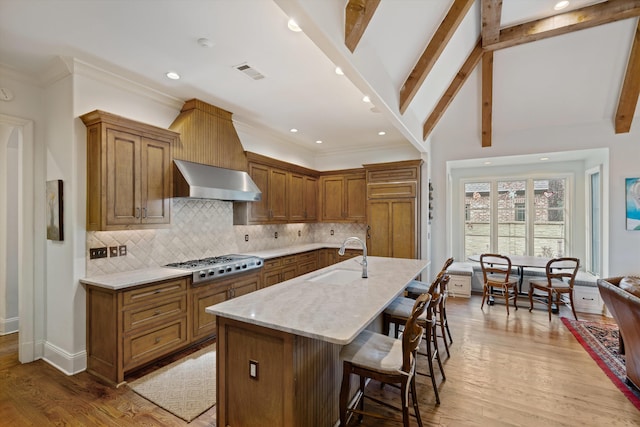 kitchen with stainless steel gas stovetop, an island with sink, a kitchen breakfast bar, wall chimney range hood, and sink