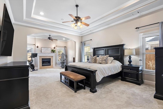 carpeted bedroom featuring multiple windows, ornamental molding, ceiling fan, a tray ceiling, and a tiled fireplace