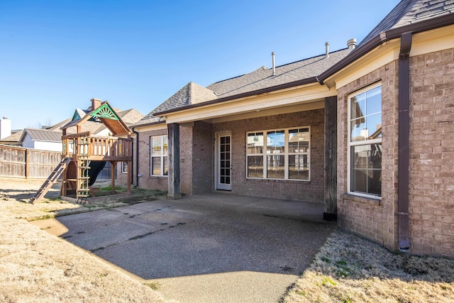 rear view of property featuring roof with shingles, a playground, a patio, brick siding, and fence