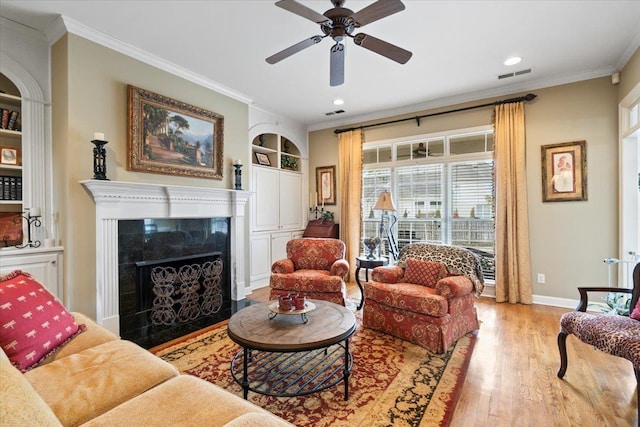 living room with ceiling fan, light wood-type flooring, a fireplace, crown molding, and built in shelves