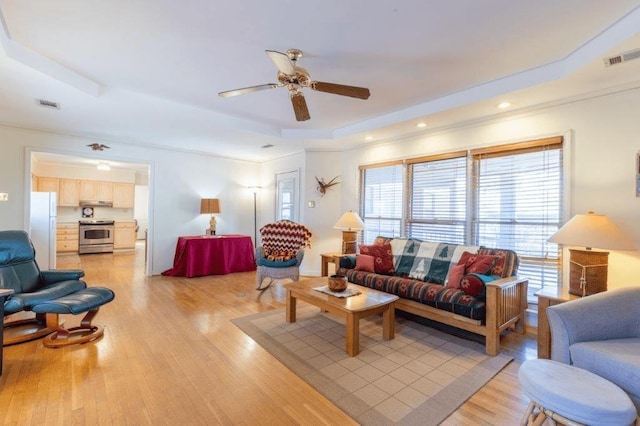 living room featuring light wood-type flooring, ceiling fan, ornamental molding, and a tray ceiling