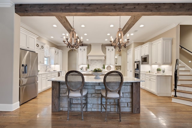 kitchen with white cabinets, appliances with stainless steel finishes, hanging light fixtures, a center island with sink, and custom range hood