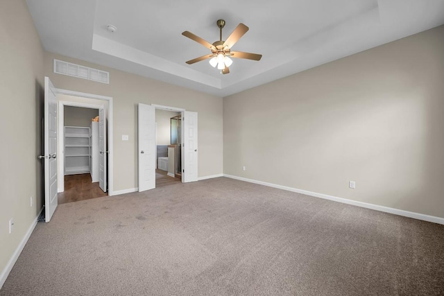 unfurnished bedroom featuring a spacious closet, a closet, a raised ceiling, ceiling fan, and dark carpet