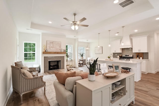 living room with sink, a tiled fireplace, ceiling fan, a tray ceiling, and light hardwood / wood-style flooring