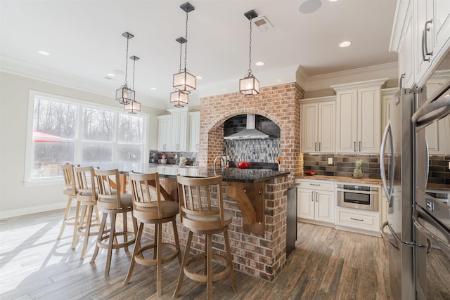 kitchen featuring a breakfast bar, decorative light fixtures, ornamental molding, a center island, and wall chimney range hood