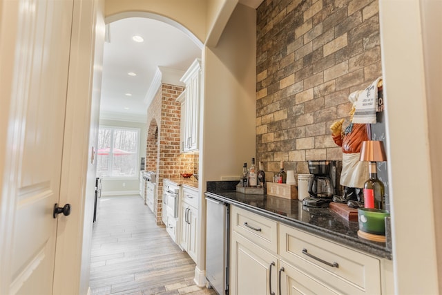 bar featuring brick wall, dark stone countertops, beverage cooler, crown molding, and light wood-type flooring