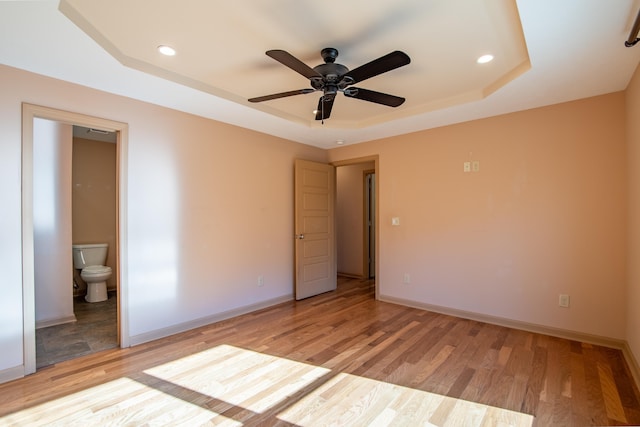 unfurnished bedroom featuring ceiling fan, light hardwood / wood-style flooring, ensuite bath, and a tray ceiling