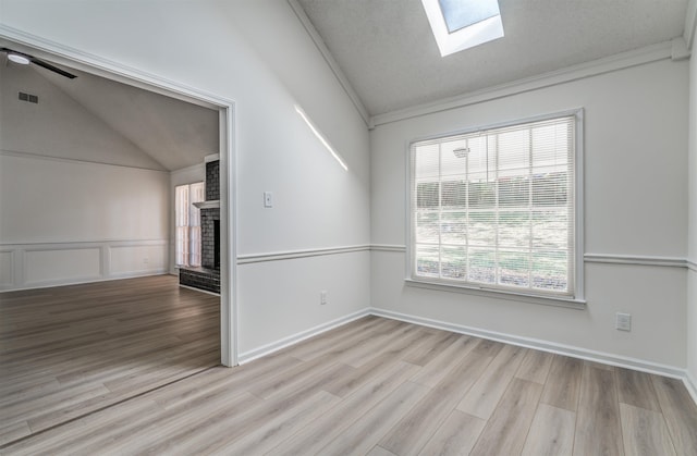 unfurnished room with crown molding, ceiling fan, vaulted ceiling with skylight, light hardwood / wood-style floors, and a brick fireplace