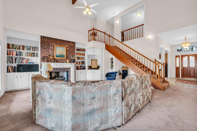 living room featuring ceiling fan with notable chandelier, a towering ceiling, a fireplace, built in shelves, and light colored carpet