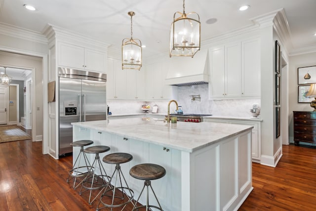 kitchen with stainless steel built in refrigerator, light stone countertops, an island with sink, white cabinets, and decorative light fixtures