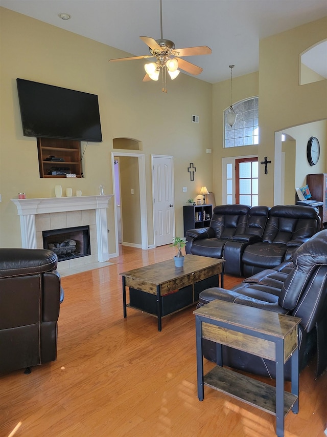 living room with ceiling fan, a tiled fireplace, a high ceiling, and light wood-type flooring