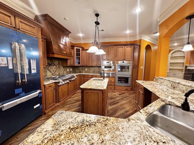 kitchen with sink, a center island with sink, pendant lighting, stainless steel appliances, and light stone countertops