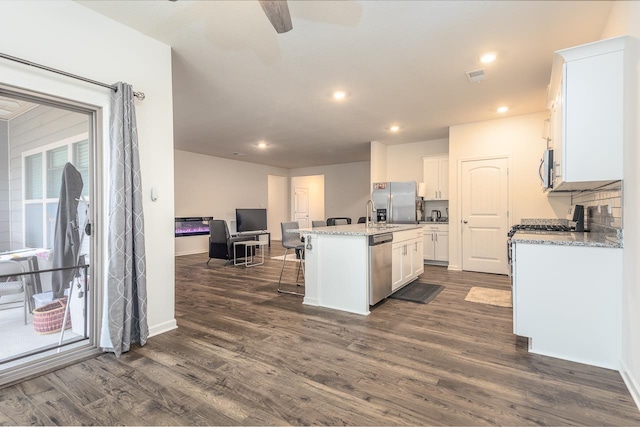 kitchen with white cabinets, dark hardwood / wood-style flooring, light stone counters, stainless steel appliances, and a center island with sink