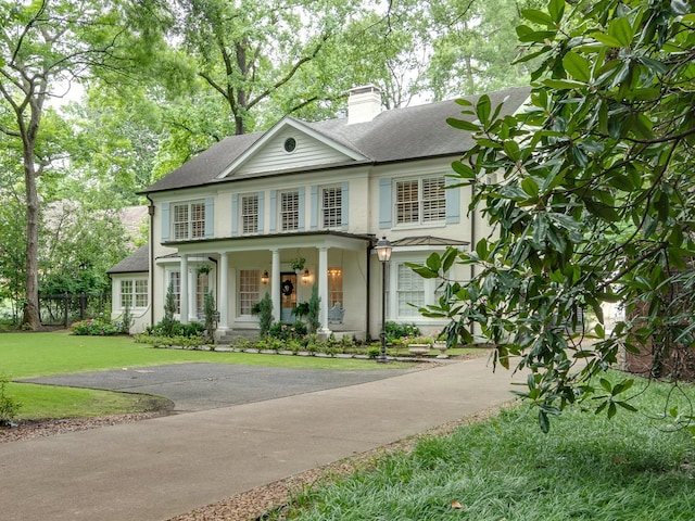 neoclassical home featuring aphalt driveway, a chimney, a porch, a shingled roof, and a front yard