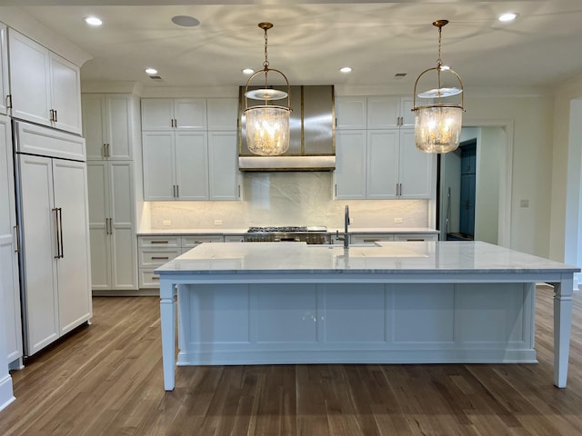 kitchen with white cabinetry, hanging light fixtures, a large island with sink, paneled built in fridge, and wall chimney range hood