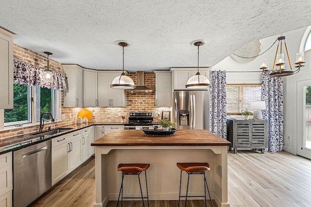 kitchen with butcher block countertops, sink, hanging light fixtures, stainless steel appliances, and wall chimney exhaust hood