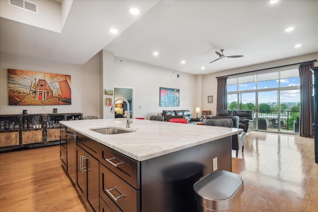 kitchen featuring sink, ceiling fan, light stone counters, an island with sink, and light wood-type flooring